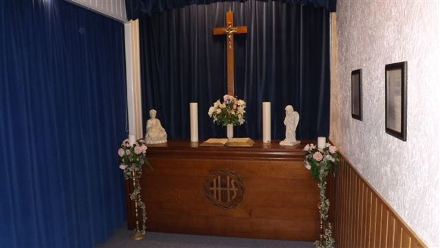 Brown & white funeral room with blue curtains, brown table, candles, book & cross